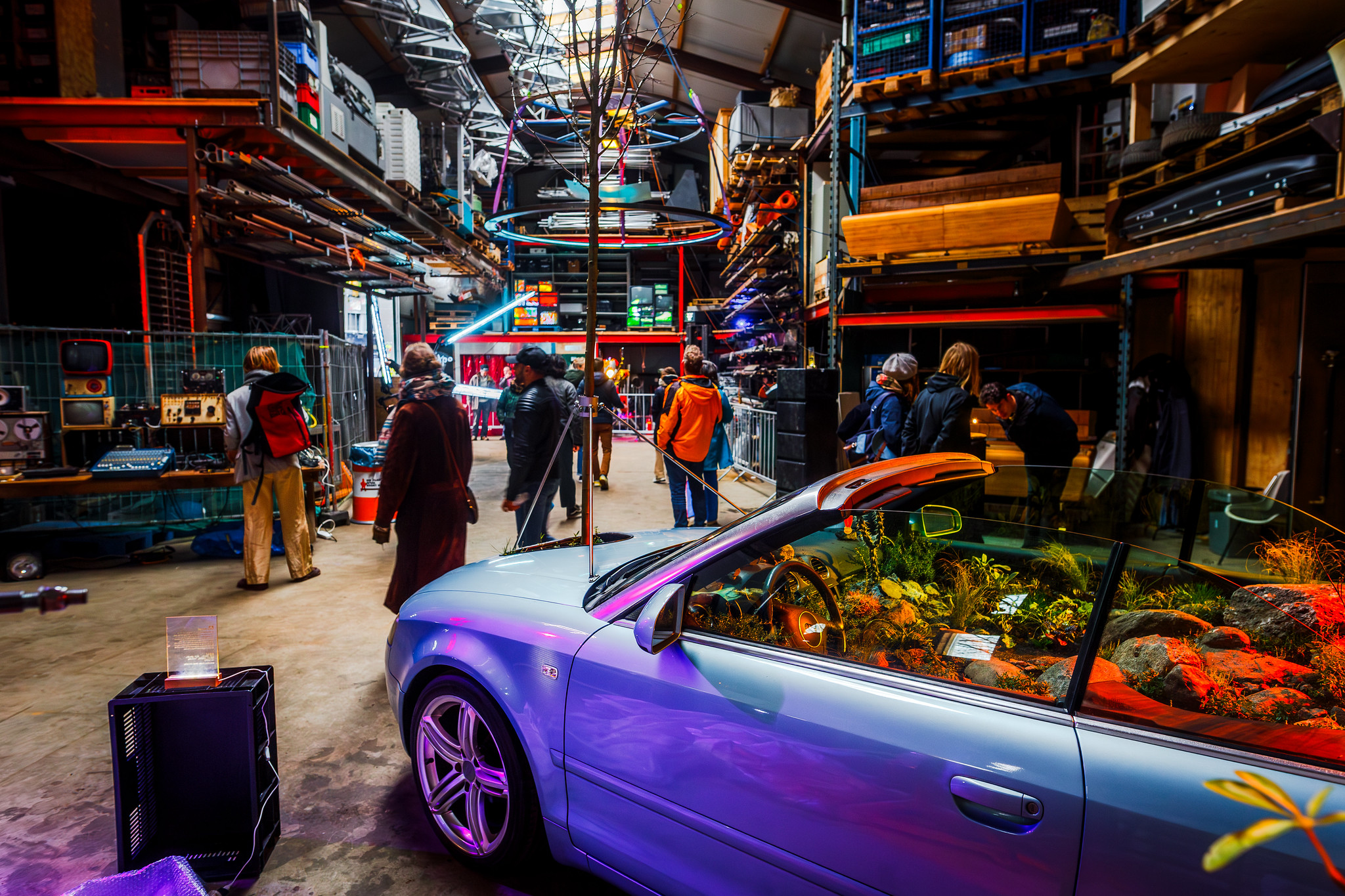 illuminated warehouse during an exposition with art installations. one of the installations in the front of the photo is convertible car filled with plants